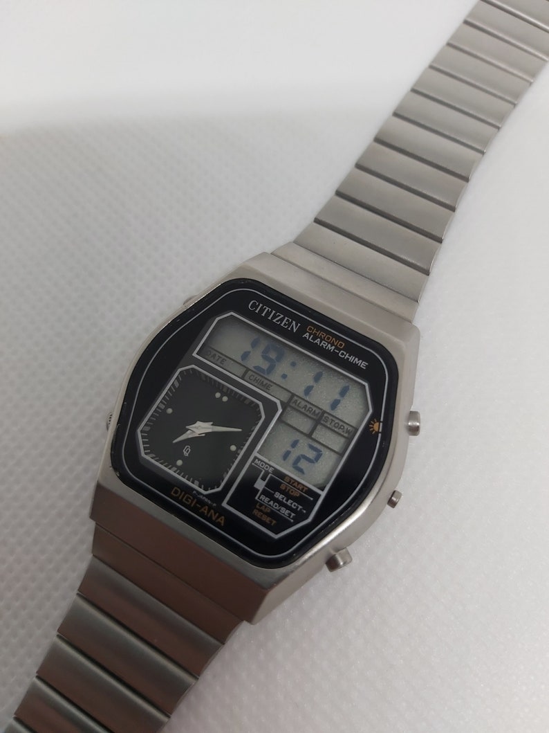 May include: A silver Citizen watch with a black face and a digital display. The watch has a chronograph, alarm, and chime functions. The watch face also has a small analog display with a black background and silver hands.