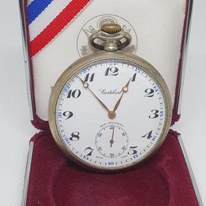 May include: A vintage Cortébert pocket watch with a white face, black numerals, and gold hands. The watch is in a silver case and displayed in a red velvet lined box with a white and red, white, and blue ribbon.
