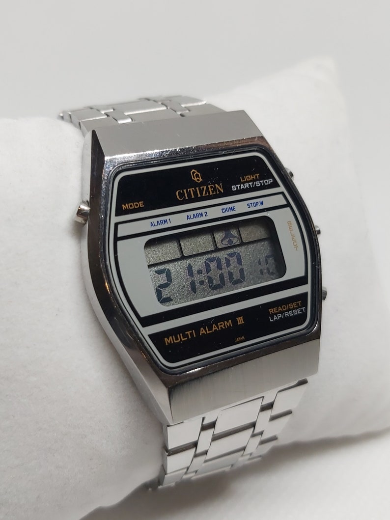 May include: A silver Citizen Multi Alarm III digital watch with a black face and a silver metal strap. The watch has a digital display showing the time as 2:00. The watch has buttons for mode, alarm 1, alarm 2, chime, stop/w, light, start/stop, read/set, and lap/reset.