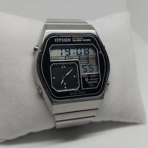 May include: A silver Citizen watch with a black face and a digital display. The watch has a chronograph, alarm, and chime functions. The watch face displays the time, date, and alarm settings. The watch has a silver metal band.