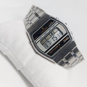 May include: A silver Citizen Multi Alarm II wristwatch with a black digital display showing the time 17:15:30. The watch has a silver metal strap and features buttons for light, start/stop, alarm, chime, and stopwatch.