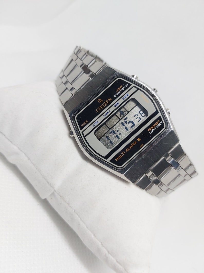 May include: A silver Citizen Multi Alarm II wristwatch with a black digital display showing the time 17:15:30. The watch has a silver metal strap and features buttons for light, start/stop, alarm, chime, and stopwatch.