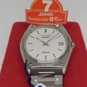 May include: A silver Citizen Seven Quartz watch with a white face and a silver metal strap. The watch has a date window at the 3 o'clock position and the words "Citizen Quartz" and "Seven" printed on the face. The watch is displayed on a red background with a red and white tag that says "Citizen Seven Quartz Battery-Life 8 Years".