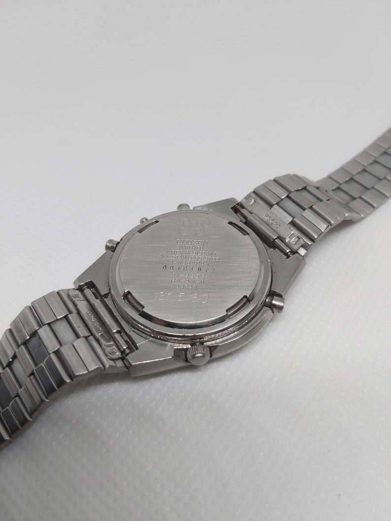 May include: A silver Citizen watch with a stainless steel band. The watch face is not visible, but the back of the watch has the Citizen logo and model number.