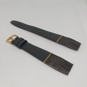 Könnte beinhalten: Zweiteiliges Uhrenarmband-Set. Die Armbänder sind dunkelgrau mit einem strukturierten, reptilienartigen Muster an den Enden. Goldfarbene Metallakzente und eine goldfarbene Schnalle vervollständigen das Design. Die Armbänder werden auf einer weißen Oberfläche präsentiert.