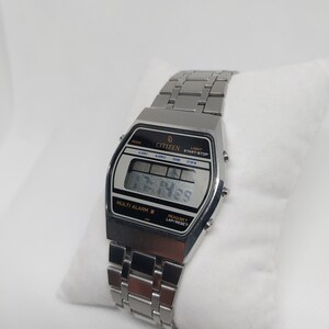 May include: A silver Citizen digital watch with a black face and a silver metal strap. The watch has a multi-alarm feature and a lap/reset function. The watch face displays the time, date, and day of the week.
