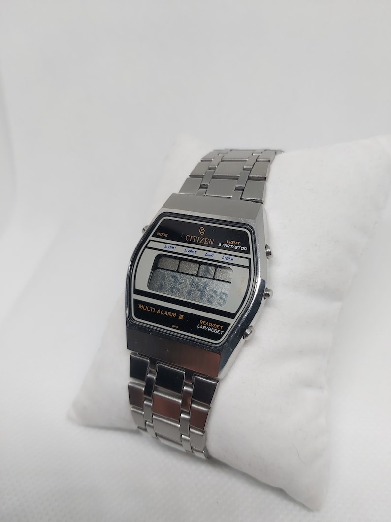 May include: A silver Citizen digital watch with a black face and a silver metal strap. The watch has a multi-alarm feature and a lap/reset function. The watch face displays the time, date, and day of the week.