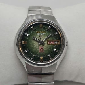 May include: A silver Seiko Elnix wristwatch with a green face and a silver metal band. The watch face has a day and date window, and the text "SEIKO ELNIX" is printed on the face.