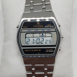 May include: A silver Citizen Multi Alarm III digital watch with a black face and a silver metal strap. The watch features a digital display showing the time, date, and alarm settings. The watch also has a range of functions, including a stopwatch, timer, and alarm.