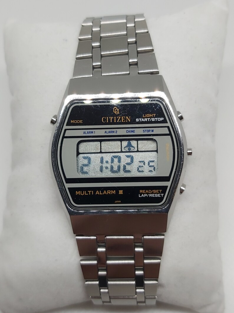 May include: A silver Citizen Multi Alarm III digital watch with a black face and a silver metal strap. The watch features a digital display showing the time, date, and alarm settings. The watch also has a range of functions, including a stopwatch, timer, and alarm.
