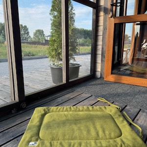 May include: A green rectangular dog bed with a brown border and the word "BUBA" printed on the bottom left corner. The bed is on a wooden floor in front of a glass door.