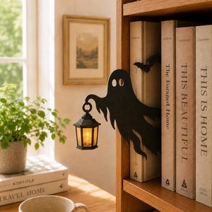 Pode incluir: Um suporte para livros em forma de fantasma preto com uma pequena lanterna suspensa que emite uma luz amarela quente. A figura do fantasma está posicionada entre livros numa prateleira de madeira, com uma silhueta de morcego preta.