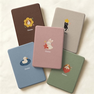 May include: Five rectangular e-reader cases in brown, beige, pink, blue, and green. Each case features a unique cartoon illustration of an animal, such as a rabbit, cat, or duck, and the name Emma printed in white text below the graphic.