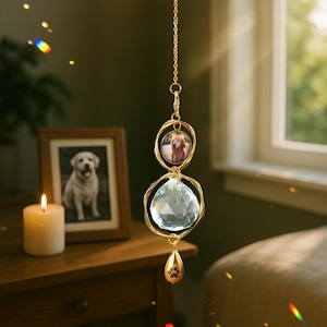 May include: A gold-colored hanging ornament featuring a clear crystal ball and a small photo of a dog. The ornament has a paw print charm at the bottom and is suspended by a delicate chain. A framed photo of a dog and a lit candle are in the background.