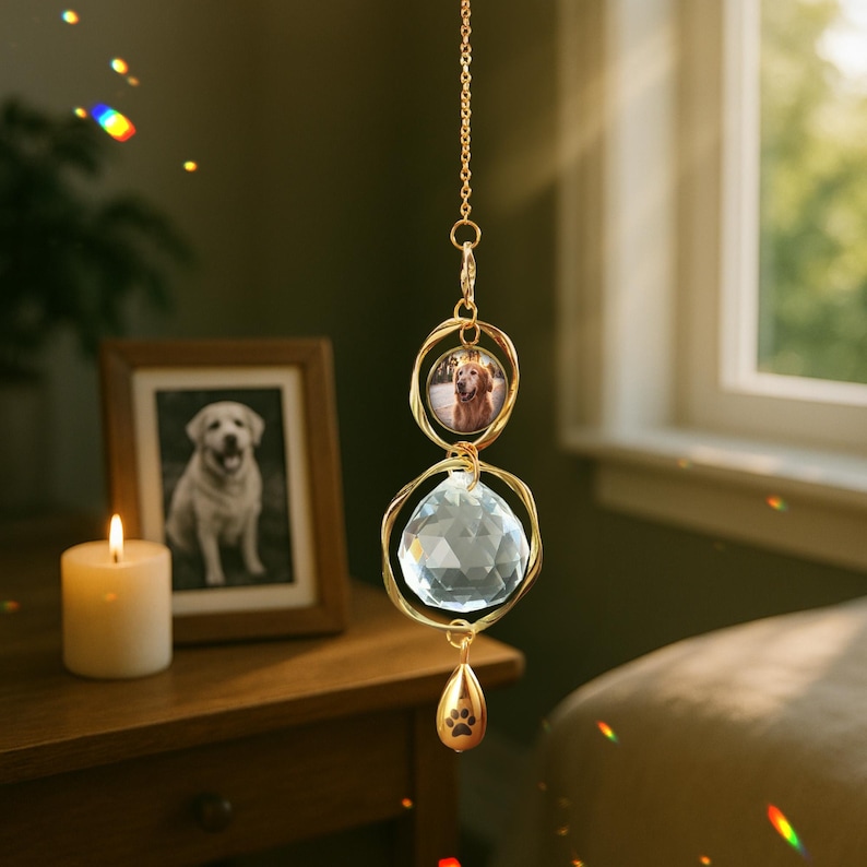 May include: A gold-colored hanging ornament featuring a clear crystal ball and a small photo of a dog. The ornament has a paw print charm at the bottom and is suspended by a delicate chain. A framed photo of a dog and a lit candle are in the background.