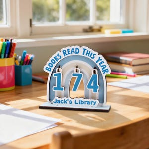May include: A wooden desk decoration with the text "BOOKS READ THIS YEAR" and the number "174" in blue. The decoration has a rainbow design and the text "Jack's Library" at the bottom. The background includes books and pens.