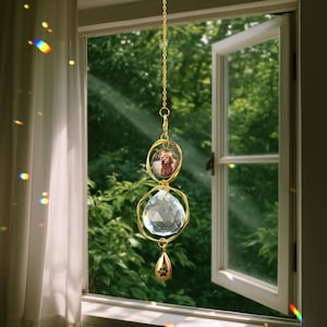 May include: A gold-colored suncatcher hanging in front of a window. The suncatcher features a photo of a dog, a large crystal ball, and a paw print charm. The background shows a green, leafy outdoor scene.
