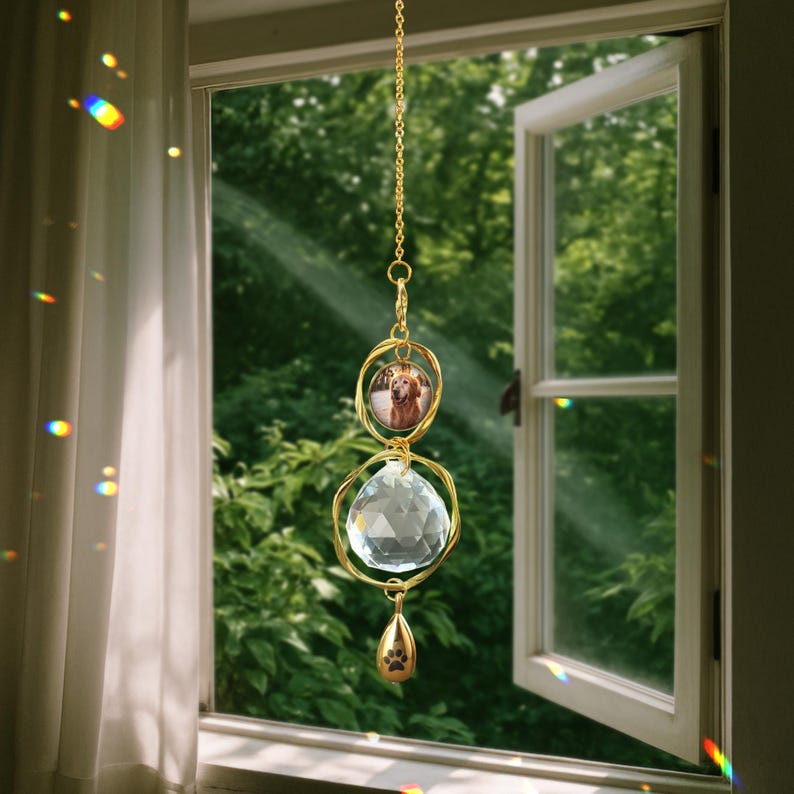 May include: A gold-colored suncatcher hanging in front of a window. The suncatcher features a photo of a dog, a large crystal ball, and a paw print charm. The background shows a green, leafy outdoor scene.