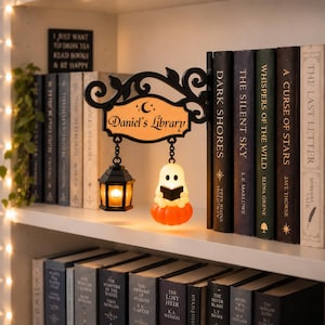 May include: A black decorative bookend featuring a sign that reads Daniel's Library with a crescent moon symbol. The sign hangs a small black lantern and a white ghost figure holding a black book, both illuminated with a warm orange glow.