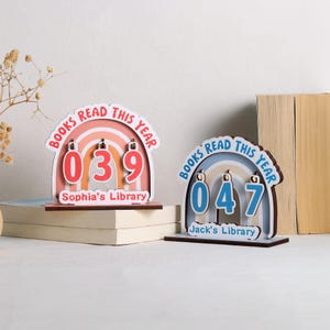 Könnte beinhalten: Zwei hölzerne Bücherzähler mit Regenbogenbögen. Einer zeigt "039" und "Sophia's Library" in Rot, Orange und Weiß. Der andere zeigt "047" und "Jack's Library" in Blau und Weiß. Beide lesen "Books Read This Year".