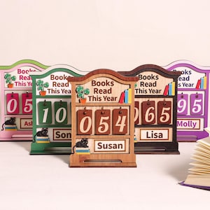 May include: A selection of wooden book counters in green, brown, and purple. Each counter displays the number of books read this year, with flip-style number blocks. The counters also feature a cat illustration, the words "Books Read This Year," and a name.