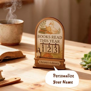 May include: Wooden desk calendar with a duck reading a book illustration. The text reads "BOOKS READ THIS YEAR" with the numbers "123" displayed. The bottom of the calendar reads "Christy's Library". A speech bubble says "Personalize Your Name".