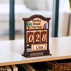 May include: A wooden book counter with the text "Books Read This Year" and the number "028". The counter has a dark brown frame with a light brown background. A small black cat and the name "Lisa" are also featured.