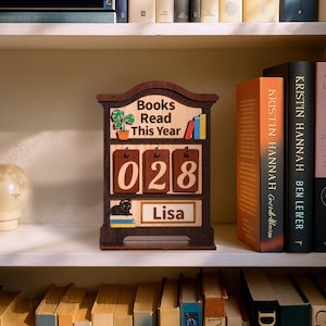May include: A wooden book tracker with the text "Books Read This Year" and the number "028". It features a small cat illustration and the name "Lisa". The tracker is displayed on a shelf with books.