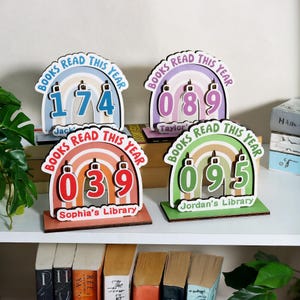 May include: Four colorful wooden book trackers with the text "BOOKS READ THIS YEAR". Each tracker displays a number and a name: 174 (Jack), 089 (Taylor), 039 (Sophia's Library), and 095 (Jordan's Library). The trackers have a rainbow design.