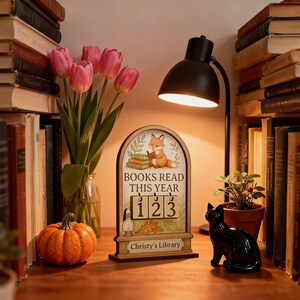 May include: A wooden desk calendar with the text "BOOKS READ THIS YEAR" and the numbers "123". The calendar features a fox reading a book, a small pumpkin, a black cat figurine, and a bouquet of pink tulips. A black desk lamp illuminates the scene.