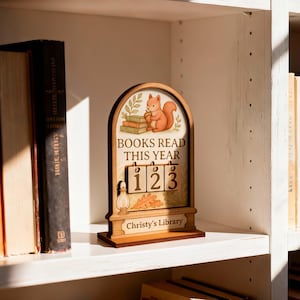 May include: Wooden bookshelf decor with a squirrel illustration above the text "BOOKS READ THIS YEAR". The counter reads "123". The base reads "Christy's Library". The decor is on a white shelf with books on either side.