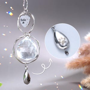 Puede incluir: Un atrapasol de cristal plateado con un dije en forma de corazón grabado con el nombre "Bella" y una huella de pata. El atrapasol tiene un pequeño dije en forma de lágrima con una huella de pata grabada.