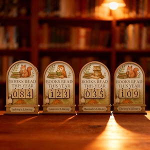 May include: Four wooden book counters with different numbers displayed. Each counter has a different animal illustration at the top, including a squirrel, owl, and raccoon. The text "BOOKS READ THIS YEAR" is above the numbers, and each counter has a name at the bottom.
