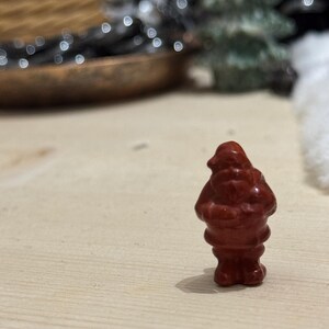 May include: A small, red figurine of Santa Claus stands on a light-colored wooden surface. The figurine has a rounded body and a detailed face. The background is blurred, with hints of a Christmas tree and decorative elements.