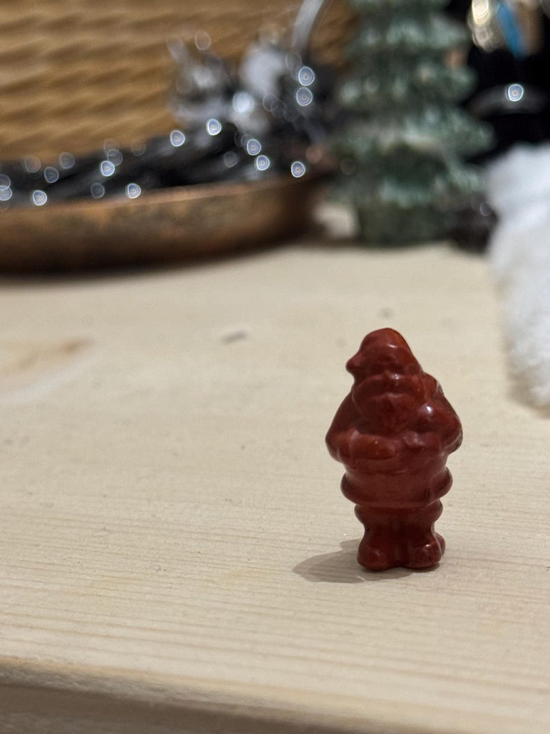 May include: A small, red figurine of Santa Claus stands on a light-colored wooden surface. The figurine has a rounded body and a detailed face. The background is blurred, with hints of a Christmas tree and decorative elements.