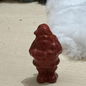 May include: A small, reddish-brown figurine of a person, possibly a gnome or a similar character. The figurine has a rounded body, a hat-like head, and a textured surface. It is standing on a light-colored surface.