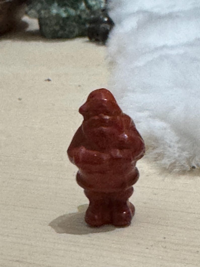 May include: A small, reddish-brown figurine of a person, possibly a gnome or a similar character. The figurine has a rounded body, a hat-like head, and a textured surface. It is standing on a light-colored surface.