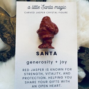 May include: A carved red jasper crystal figure of Santa Claus, displayed on a white card with text. The card reads "a little Santa magic" and "SANTA generosity + joy". The text also mentions red jasper's properties.