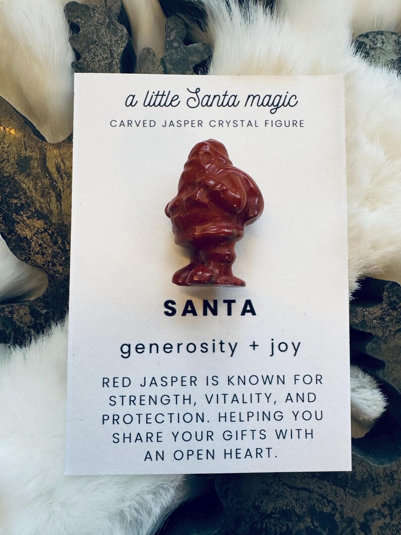 May include: A carved red jasper crystal figure of Santa Claus, displayed on a white card with text. The card reads "a little Santa magic" and "SANTA generosity + joy". The text also mentions red jasper's properties.