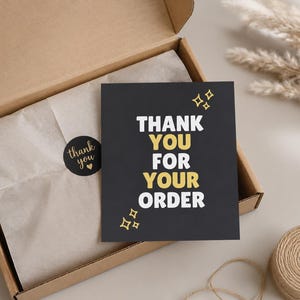 Könnte beinhalten: Eine quadratische schwarze Karte mit dem weißen und goldenen Text THANK YOU FOR YOUR ORDER und goldenen Sternsymbolen in einem offenen braunen Versandkarton. Der Karton enthält hellbraunes Seidenpapier, das mit einem runden schwarzen Aufkleber.