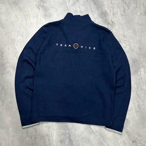 K&ouml;nnte beinhalten: Marineblaues Sweatshirt mit Stehkragen und langen &Auml;rmeln. Der Text "TEAM NIKE" ist in Wei&szlig; auf der R&uuml;ckseite aufgedruckt, zusammen mit dem Nike-Logo. Die Manschetten und der Saum sind grau eingefasst.