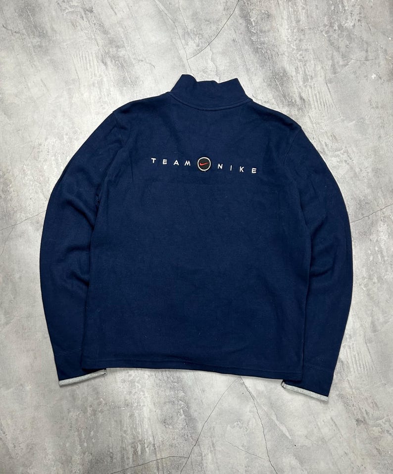 K&ouml;nnte beinhalten: Marineblaues Sweatshirt mit Stehkragen und langen &Auml;rmeln. Der Text "TEAM NIKE" ist in Wei&szlig; auf der R&uuml;ckseite aufgedruckt, zusammen mit dem Nike-Logo. Die Manschetten und der Saum sind grau eingefasst.