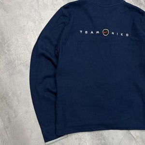 K&ouml;nnte beinhalten: Marineblaues Langarm-Sweatshirt mit Stehkragen. Der Text "TEAM NIKE" ist in Wei&szlig; auf der R&uuml;ckseite aufgedruckt, mit dem Nike-Logo in Rot und Orange. Die &Auml;rmelb&uuml;ndchen haben einen grauen Besatz.