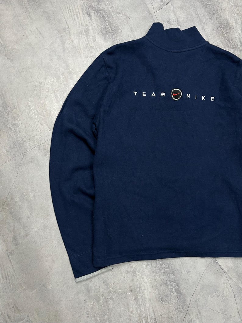 K&ouml;nnte beinhalten: Marineblaues Langarm-Sweatshirt mit Stehkragen. Der Text "TEAM NIKE" ist in Wei&szlig; auf der R&uuml;ckseite aufgedruckt, mit dem Nike-Logo in Rot und Orange. Die &Auml;rmelb&uuml;ndchen haben einen grauen Besatz.