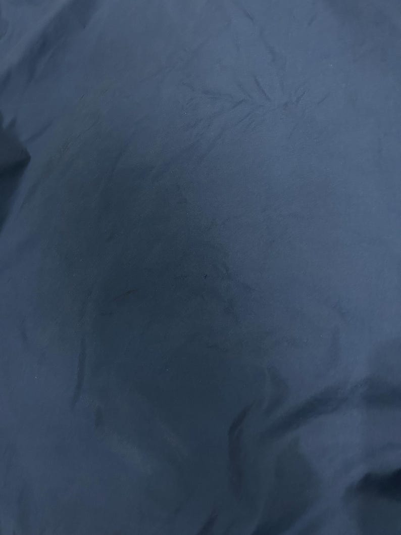 Op de afbeelding: Een close-up van een marineblauwe stof met een licht gekreukte textuur. De stof lijkt glad te zijn, mogelijk van synthetisch materiaal. De belichting cre&euml;ert subtiele highlights en schaduwen.