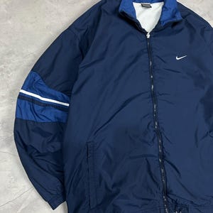 Op de afbeelding: Marineblauwe Nike-trainingsjas met een wit Nike swoosh-logo op de borst. De jas heeft een volledige ritssluiting, een opstaande kraag en een colorblock-ontwerp op de mouwen met witte strepen. De jas is gemaakt van een lichtgewicht materiaal.