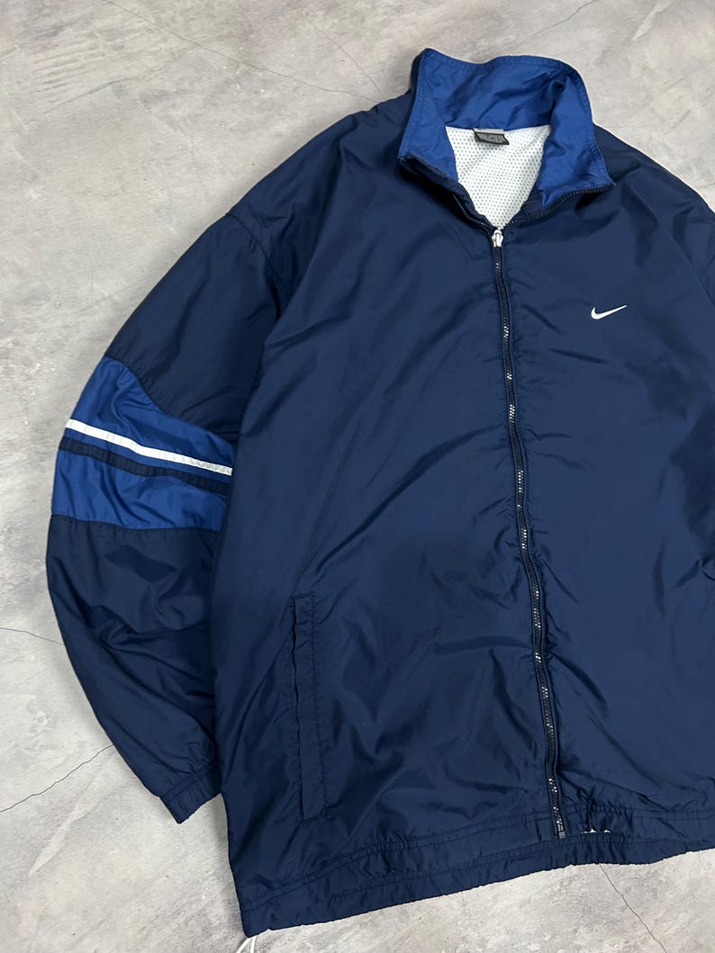 Op de afbeelding: Marineblauwe Nike-trainingsjas met een wit Nike swoosh-logo op de borst. De jas heeft een volledige ritssluiting, een opstaande kraag en een colorblock-ontwerp op de mouwen met witte strepen. De jas is gemaakt van een lichtgewicht materiaal.