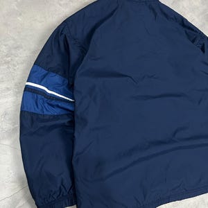 Op de afbeelding: Marineblauwe jas met een hoge kraag. De jas heeft een colorblock-ontwerp op de mouwen met donkerblauwe en witte strepen. De jas is gemaakt van een lichtgewicht materiaal.