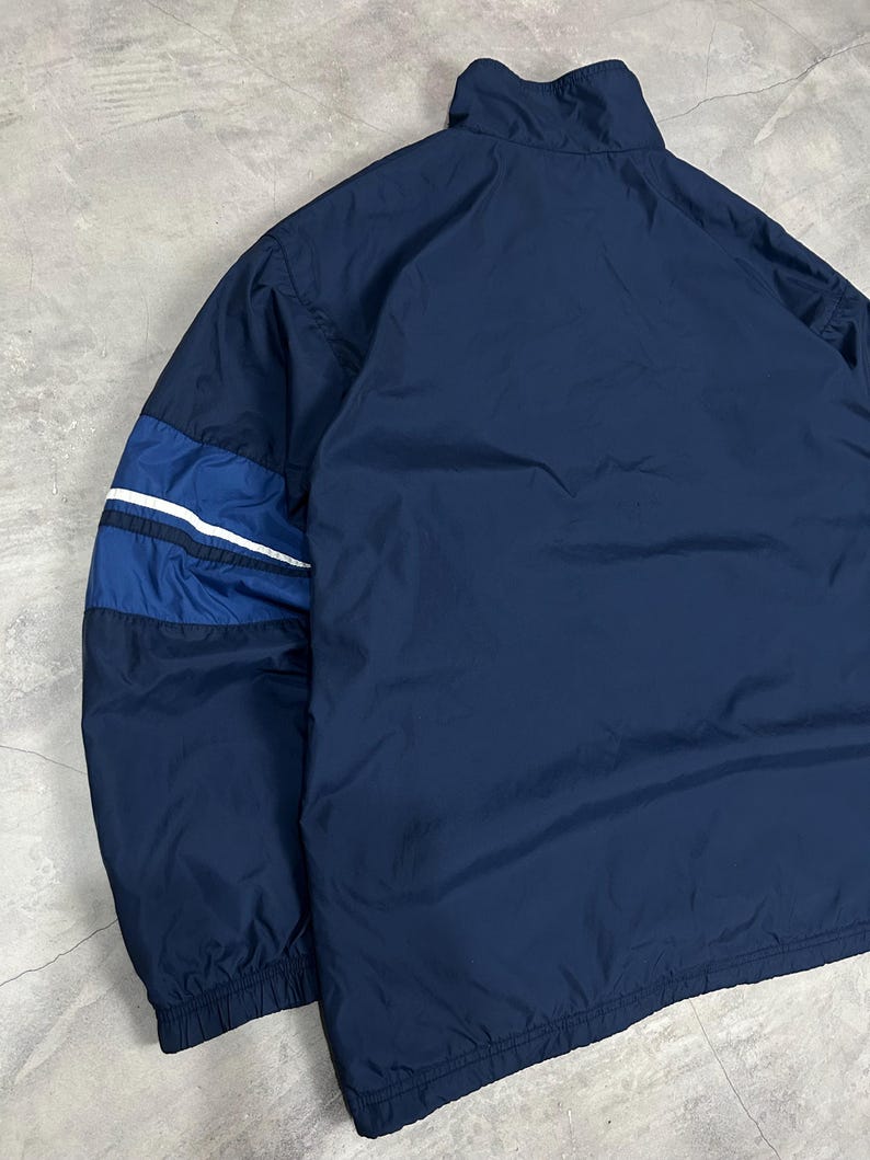 Op de afbeelding: Marineblauwe jas met een hoge kraag. De jas heeft een colorblock-ontwerp op de mouwen met donkerblauwe en witte strepen. De jas is gemaakt van een lichtgewicht materiaal.