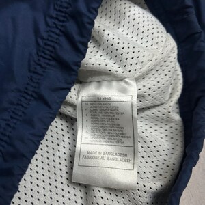 Op de afbeelding: Close-up van de binnenkant van een marineblauw kledingstuk, met een witte mesh voering. Een label toont tekst met materiaal samenstelling en "MADE IN BANGLADESH". De marineblauwe buitenkant van het kledingstuk is zichtbaar.